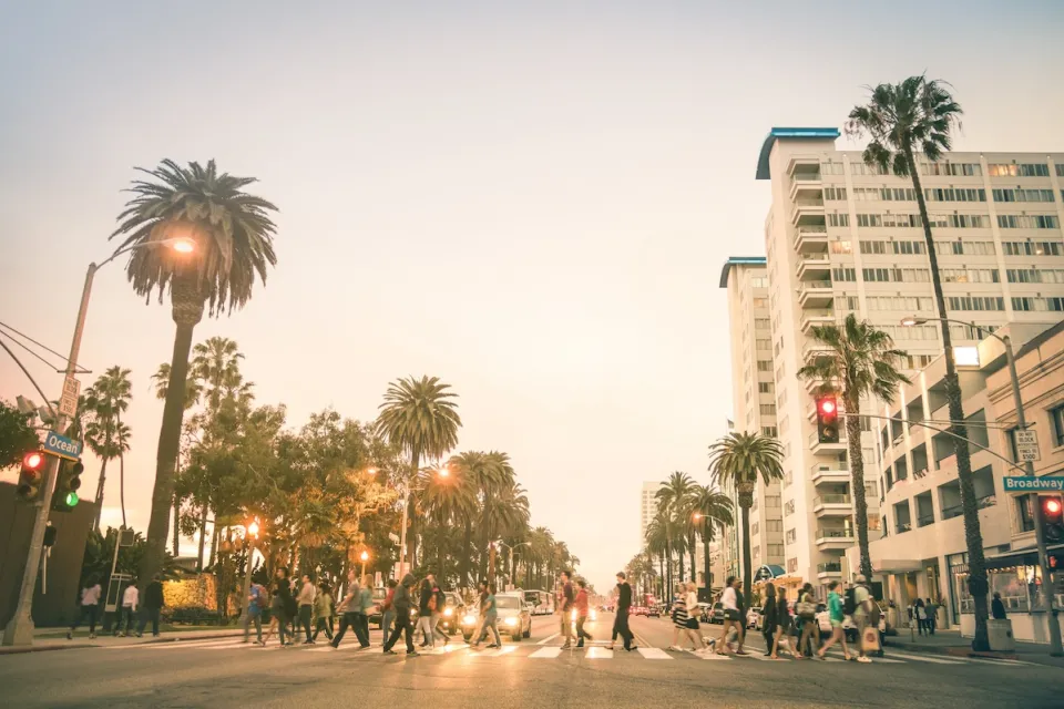 Busy pedestrian crosswalk on wide boulevard in Santa Monica, California lined with tall palm trees against golden hour light.
